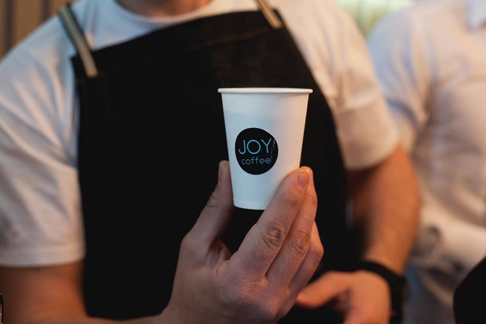 Barista cu șorț negru ținând un pahar JOY coffee