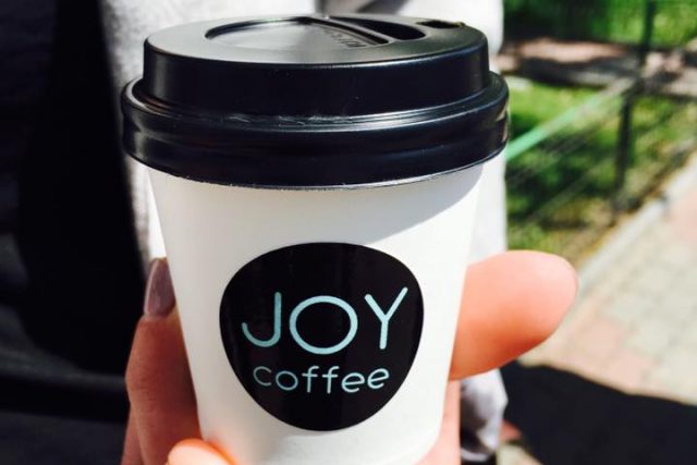 A lidded JOY coffee cup held in hand, out on the street